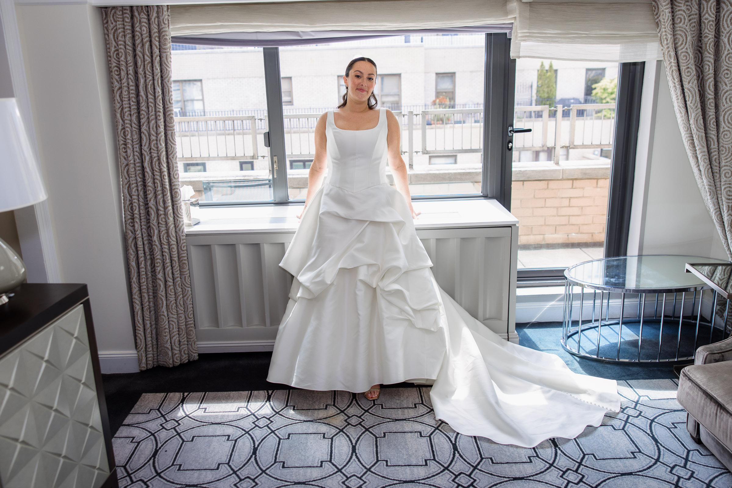 Bride in a layered white gown posing by the hotel window before the wedding
