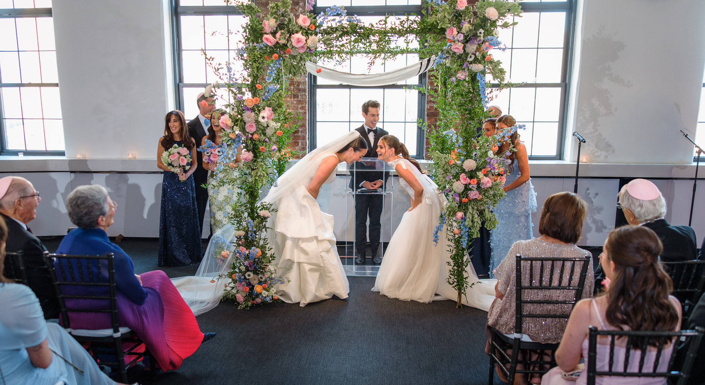 Wide view of Sydney and Delilah’s indoor ceremony framed by windows, brick walls, and colorful florals