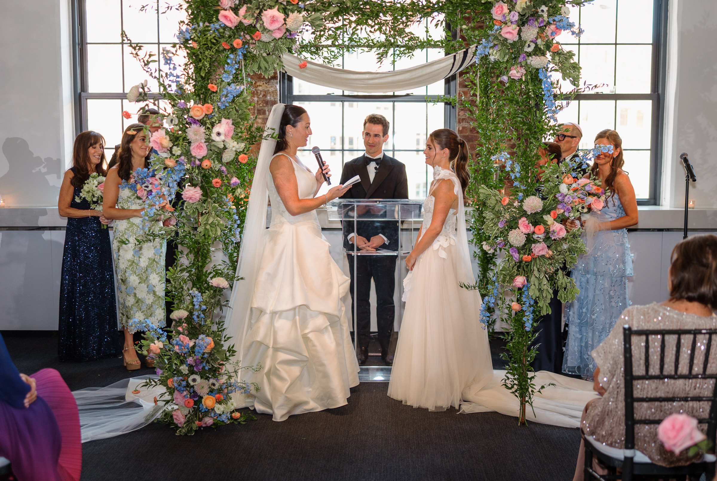 The brides exchanging vows beneath a colorful floral ceremony structure indoors